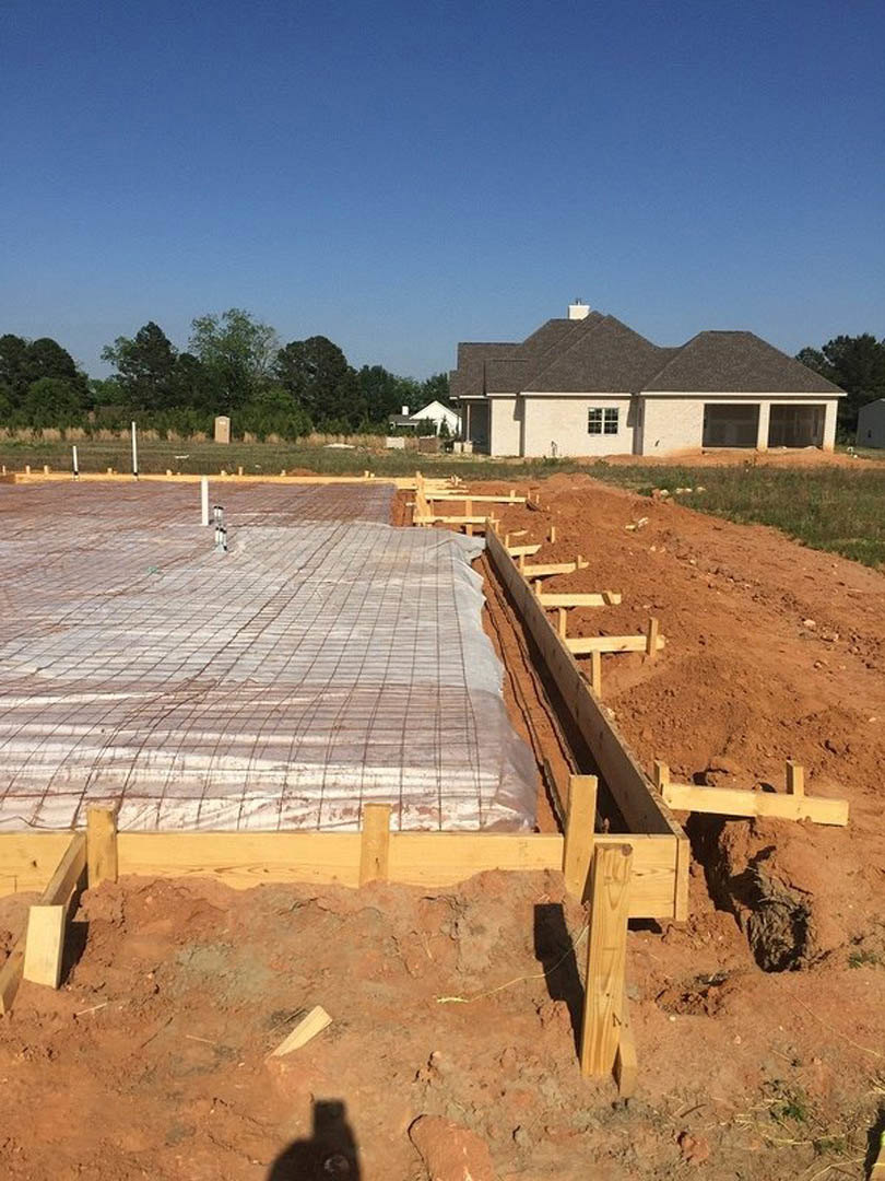 Wooden foundation framing on a construction site with exposed soil and grass, blue sky overhead, and a tree in the background.