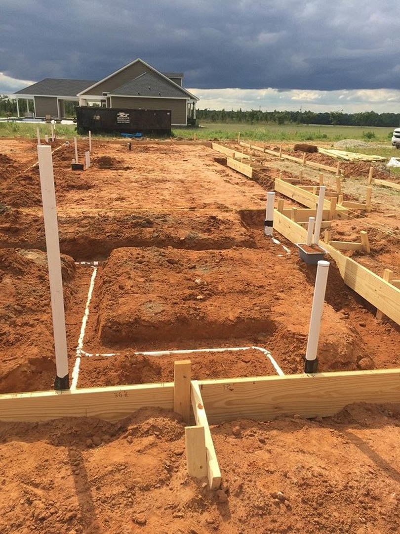 Partially built house with exposed framing, white support poles, dirt lot, and blue sky in background