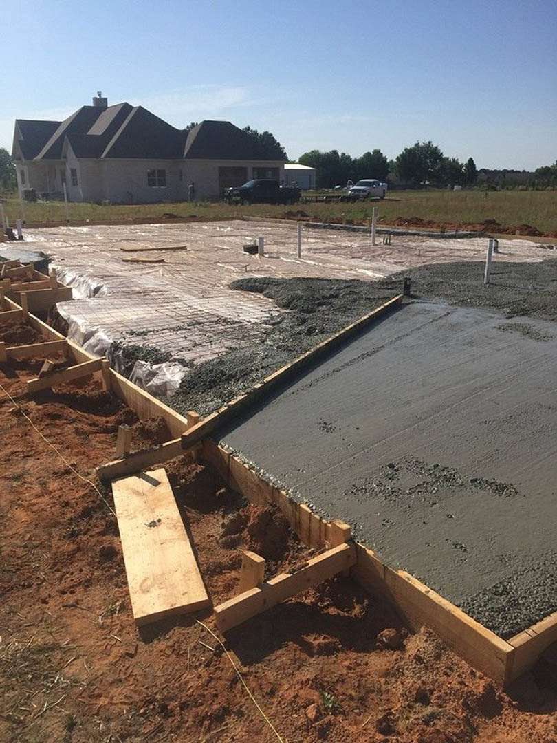 Concrete foundation and framing in progress on custom home construction site, chimney visible, wood planks scattered on ground, blue sky with scattered clouds overhead, trees in