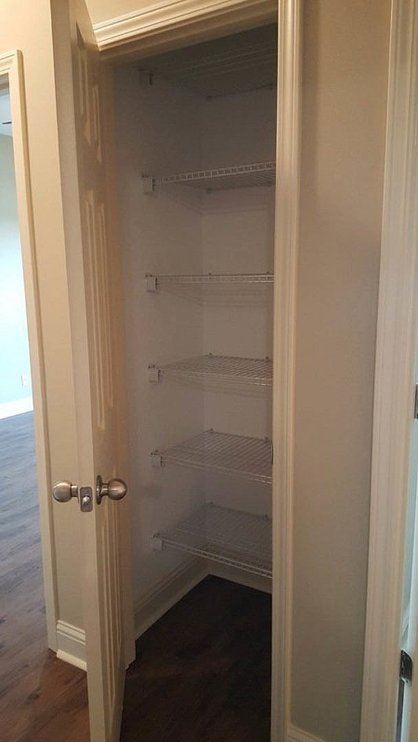 Walk-in closet with white built-in shelves, dark wood flooring, white baseboard trim, close-up of brushed metal door knob, wall vent, and smooth white plaster walls