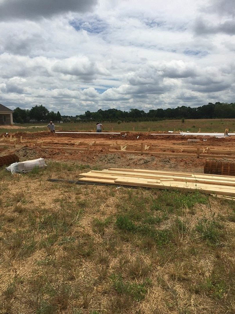 Framed house under construction with workers on grassy lot, scattered wood planks and piles, cloudy sky overhead, white animal resting nearby