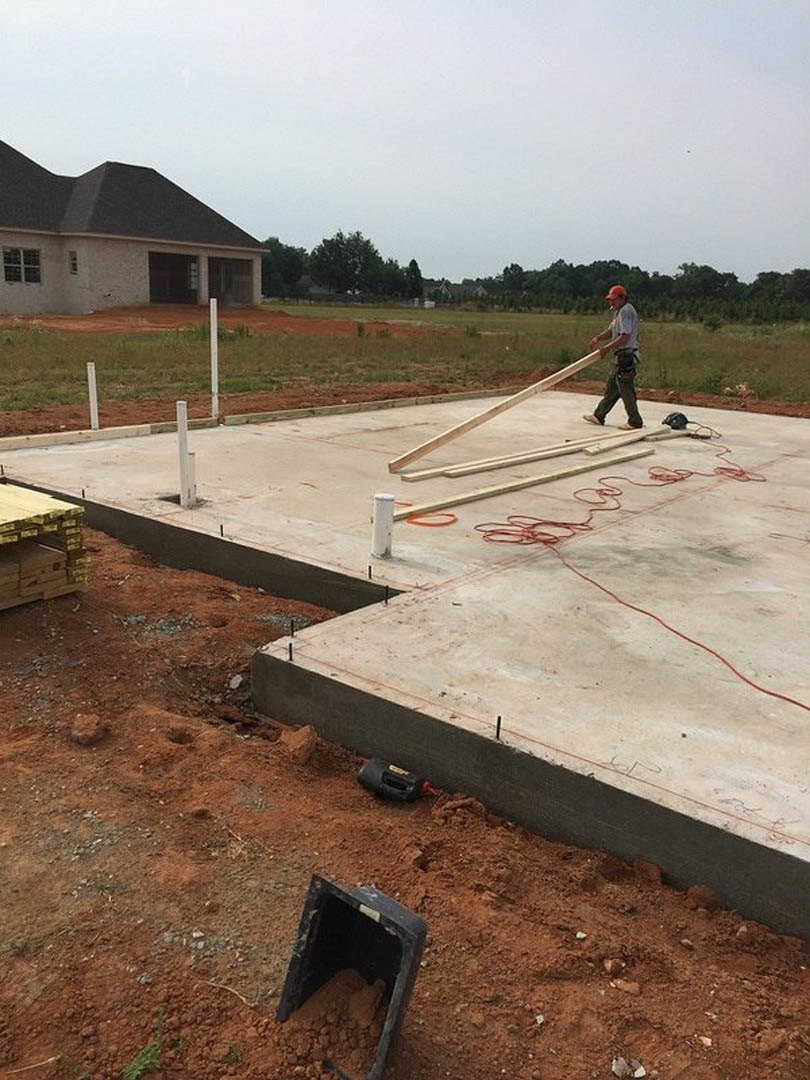 Man holding wooden beam near stack of wood planks outside custom home with black roof, dark-framed window, and grassy yard.