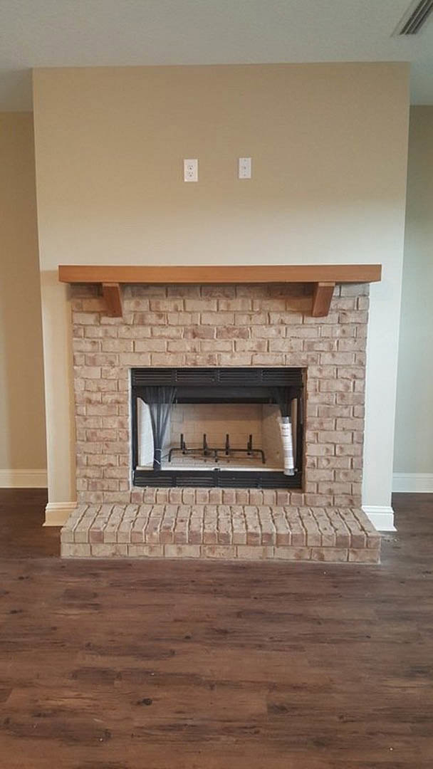 Black-framed fireplace with glass door set in a brick wall, wood mantel shelf above, wood flooring and brick steps in foreground