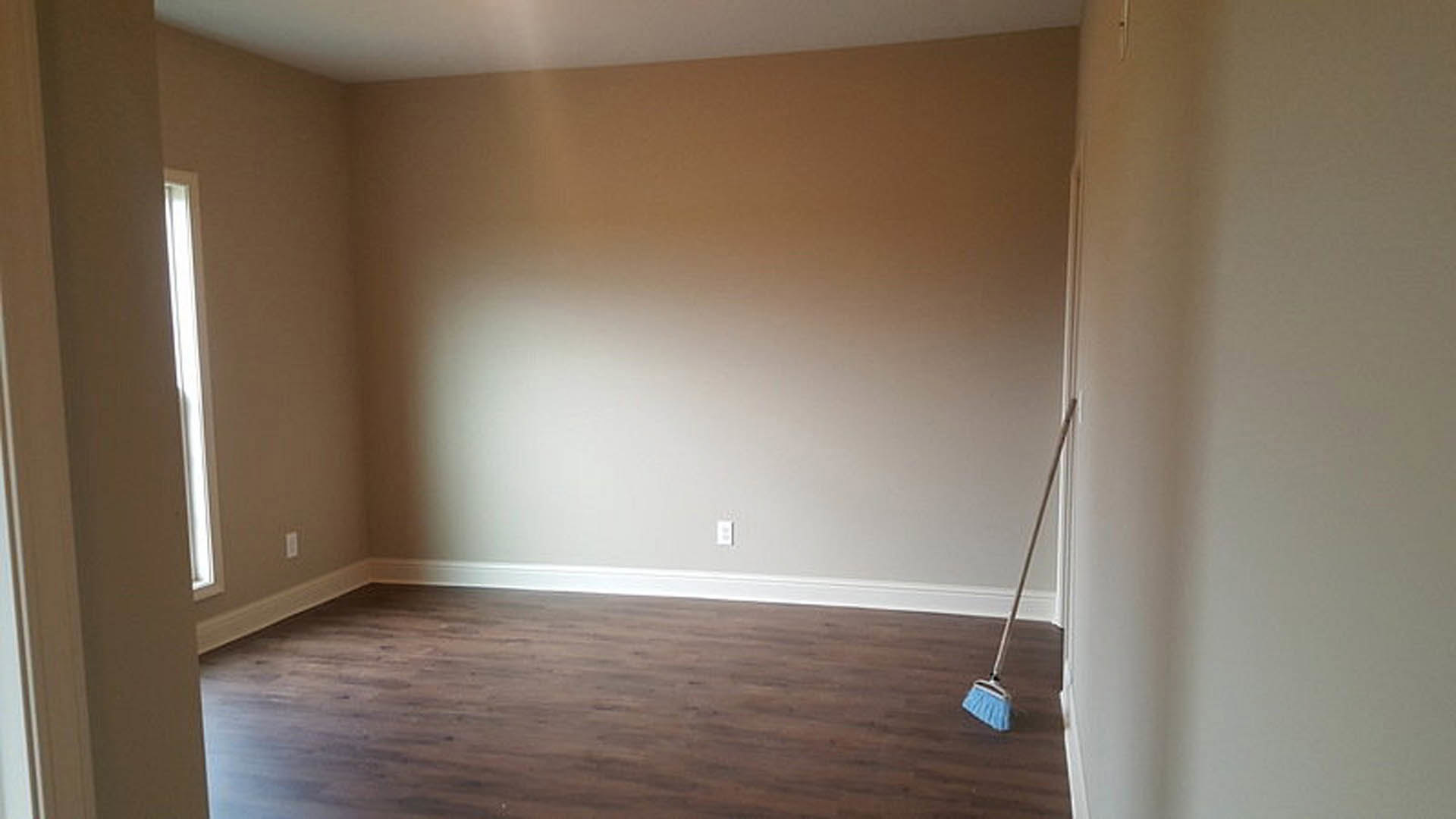 Blue dustpan and mop resting on brown laminate flooring, sunlight streaming through window onto plaster walls, wood ceiling visible above.