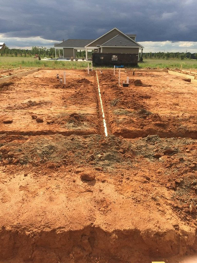 Wood-framed house under construction on a dirt lot with exposed soil and construction poles, completed home and cloudy sky in the background
