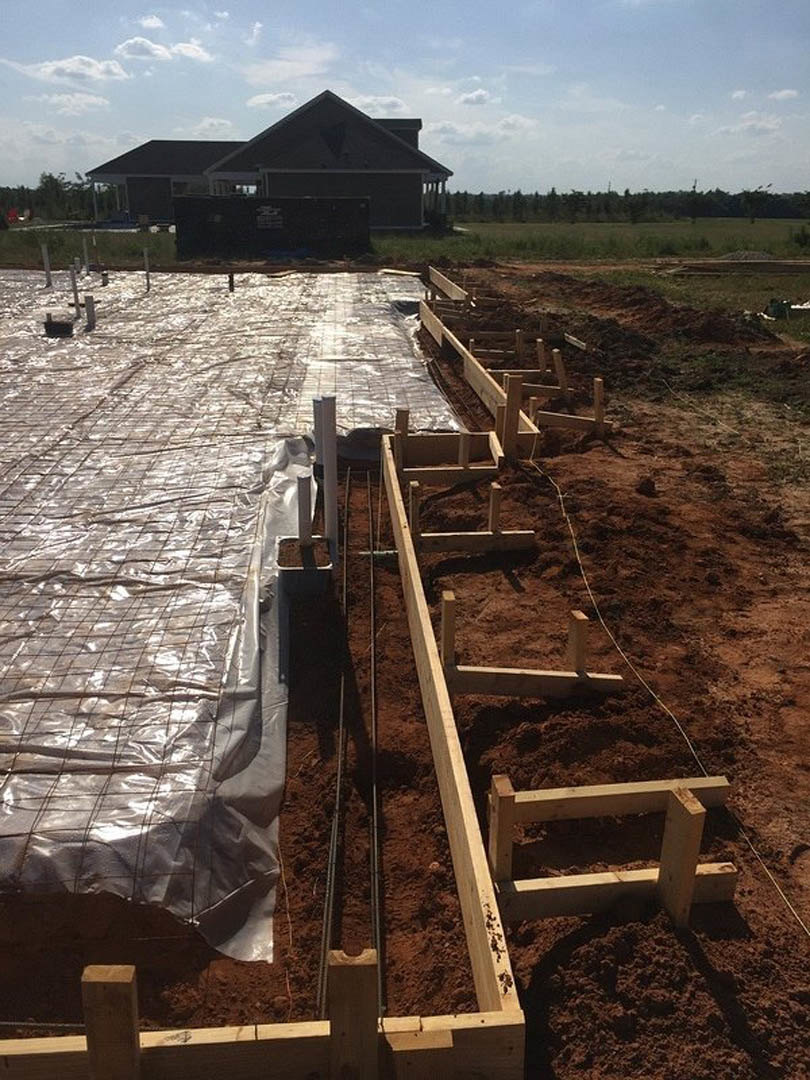 Wood-framed house under construction with exposed beams and metal bars, tarp covering part of the site, completed house with triangular roof in the background, grassy ground, blue