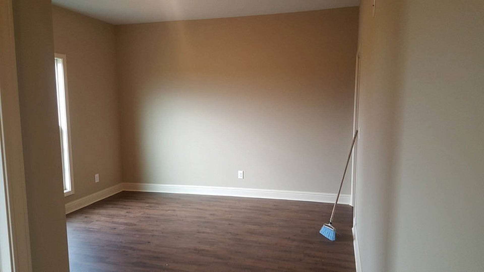 Brown wood laminate floor with white baseboard, broom and blue dustpan near wall, sunlight streaming through window onto plaster wall