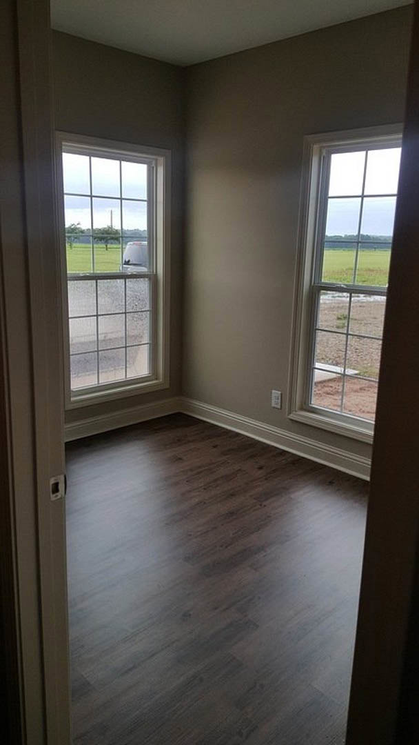 Sunlit room featuring dark wood flooring, white baseboard trim, and large windows overlooking a grassy field.