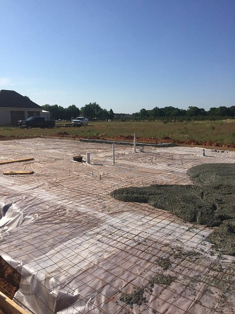 Construction site with piles of gravel, metal grids, and bars in front of a partially built house, car parked nearby, clear blue sky and trees in background