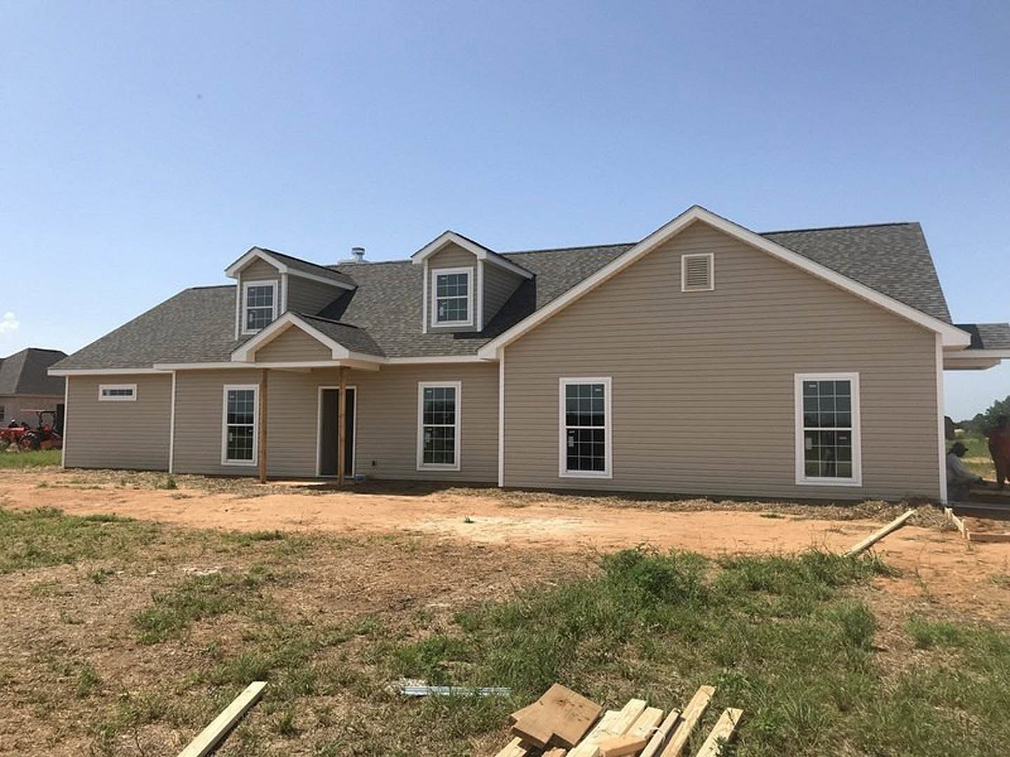 Partially built house with white-framed multi-pane windows, dormer, exterior siding, and vent, surrounded by dirt field and patchy grass