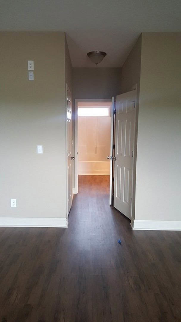 Hallway with wood flooring, white walls, two doors, light fixture, and visible light switch; open door reveals bathroom with bathtub