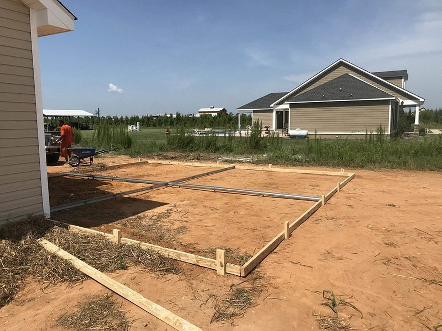 Partially built house with exposed framing and roof, construction worker in orange jumpsuit standing on dirt lot, metal pipes and construction materials scattered around, trees and
