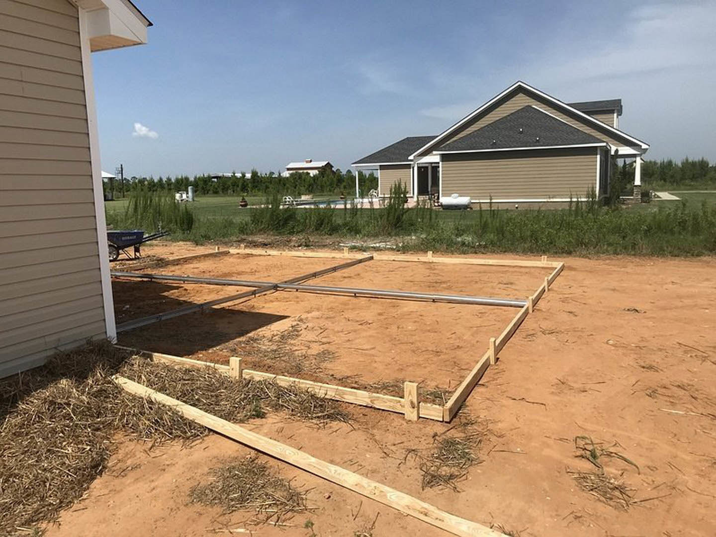 Modern house with gray roof, wooden fence enclosing yard, dirt ground with straw pile, blue wheelbarrow near entrance, surrounding grass and trees under partly cloudy sky