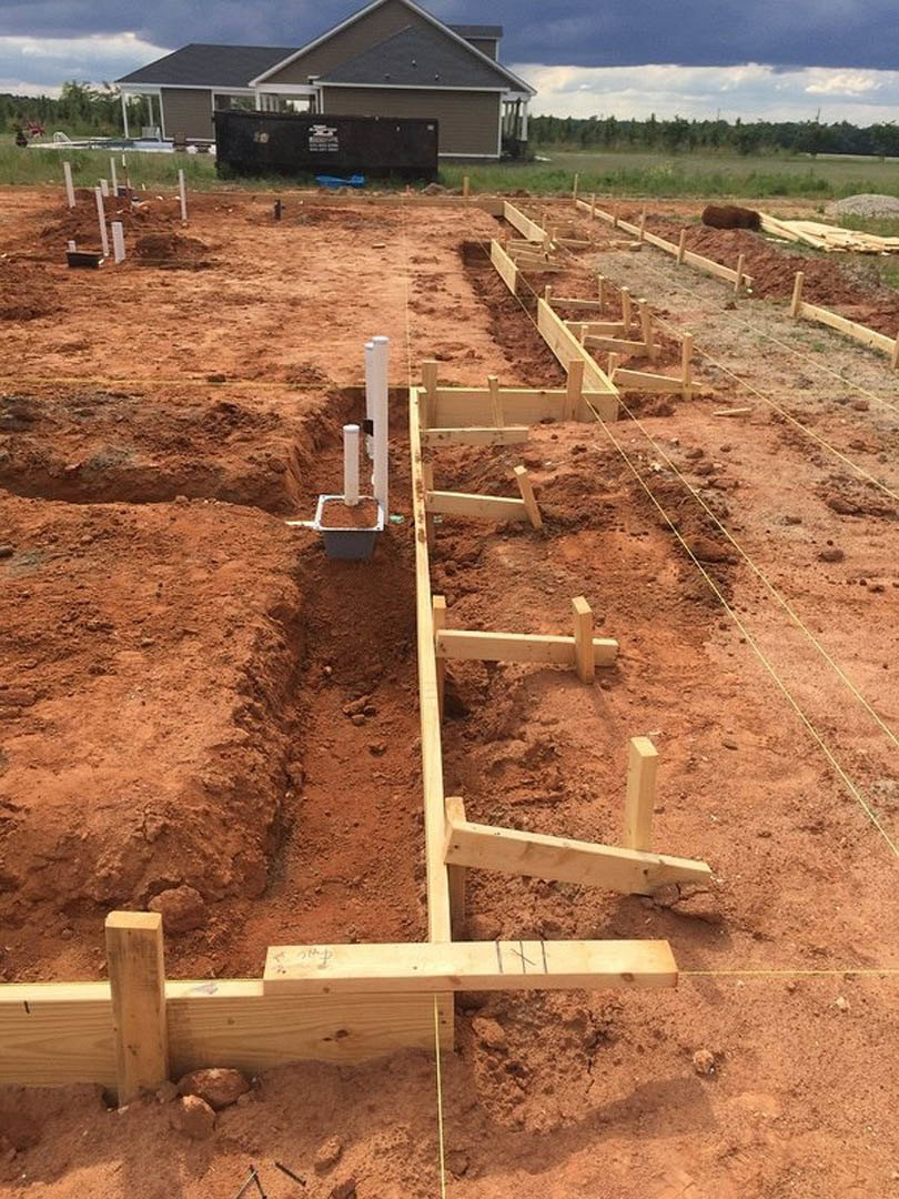 Wood-framed house under construction with exposed beams, black exterior wall, white support post, and dirt foundation surrounded by grass.