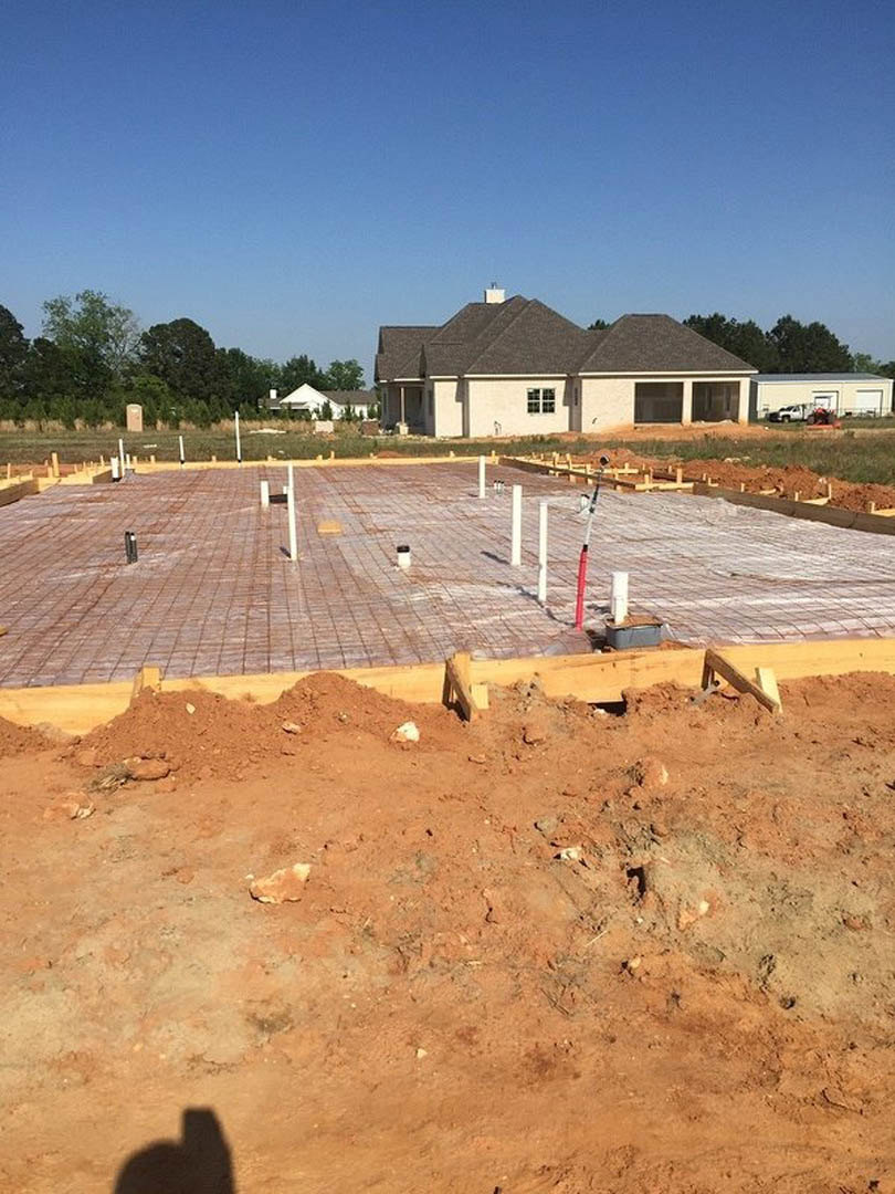 Wood framing and dirt foundation of a house under construction, partially completed roof, blue sky, and finished home in the background
