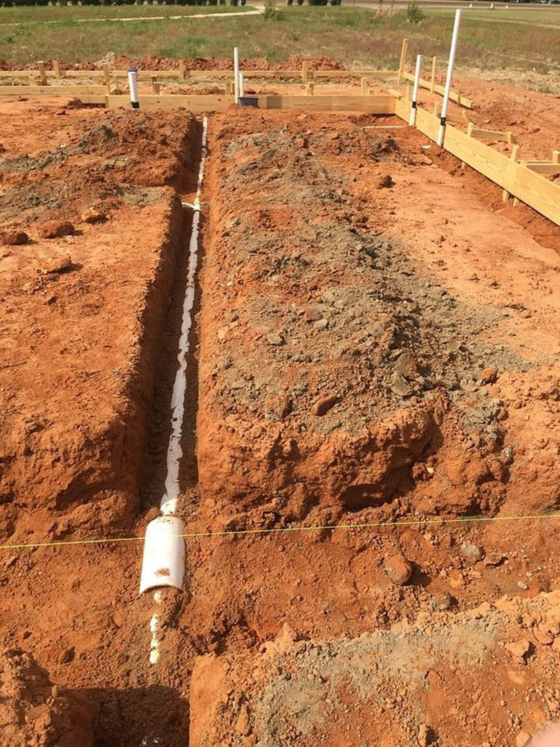 Narrow trench dug in brown soil with exposed pipe, surrounded by patches of grass and loose dirt