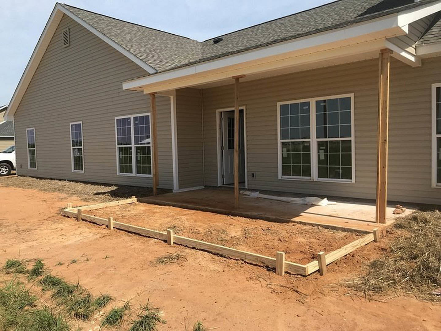 Partially built home with exposed framing, fenced dirt yard, covered porch, white door with glass window, scattered wooden planks, and light siding under a clear sky