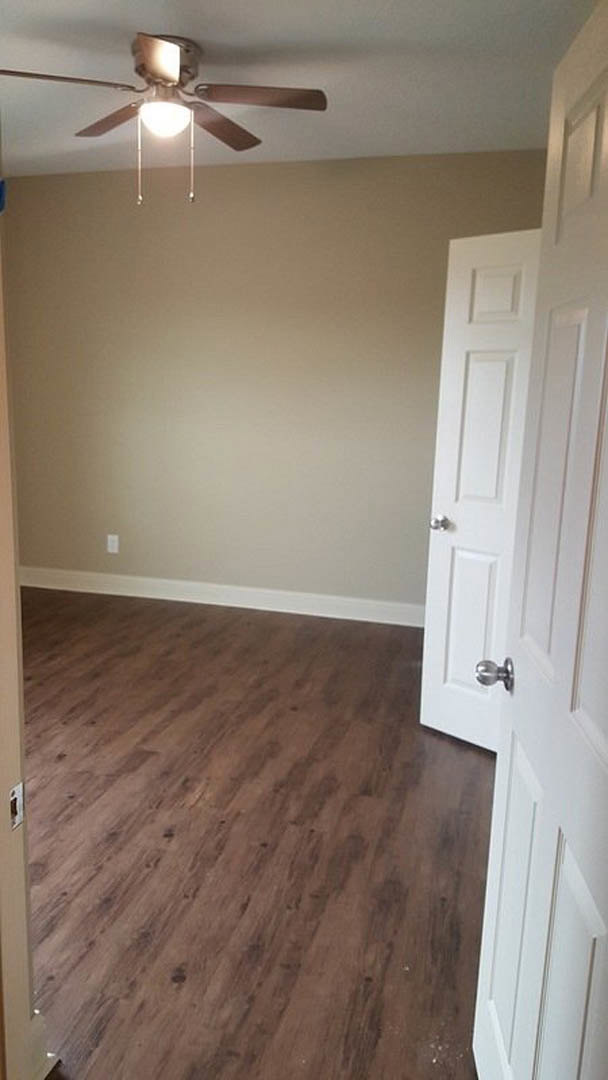 Wood flooring in a room with white paneled door, ceiling fan with light fixture, neutral walls, and simple trim.
