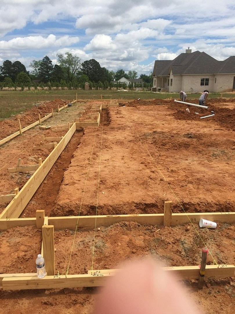 Partially built house with shingled roof, exposed foundation, workers on dirt lot, scattered construction materials, grassy patches, trees in background, cloudy sky overhead, pink