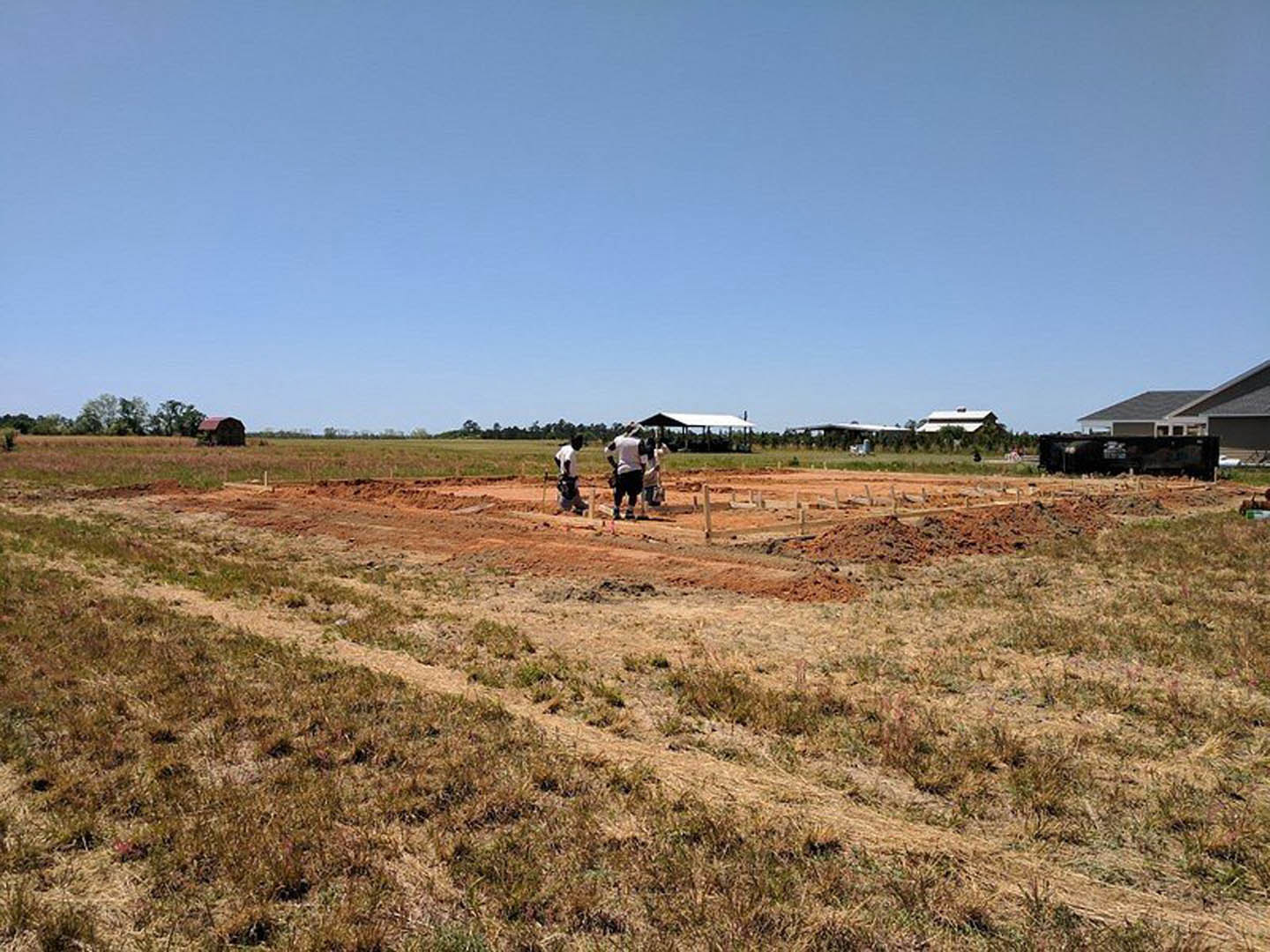 Construction crew building custom home on grassy field under clear blue sky, with exposed soil, scattered plants, and a white truck in the background