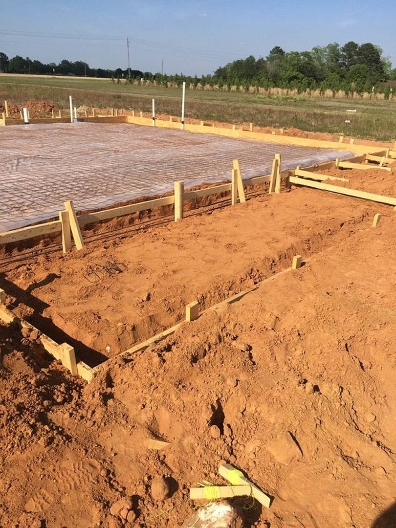 Wooden forms and dirt foundation at a residential construction site, fenced perimeter, string marking layout, blue sky and clouds, trees in background