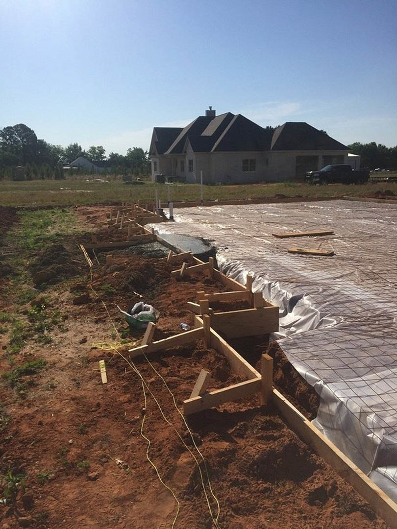 Partially constructed house with exposed wooden framing, chimney, dirt construction site, black van parked nearby, trees and grass in background, blue sky overhead