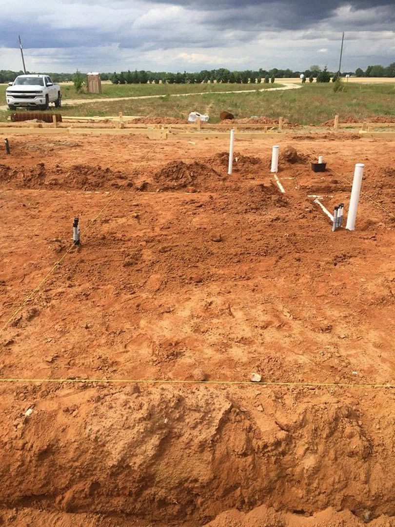 Dirt construction site with white utility poles, grassy field, scattered dirt mounds, white truck parked on road, cloudy sky overhead, trees in background