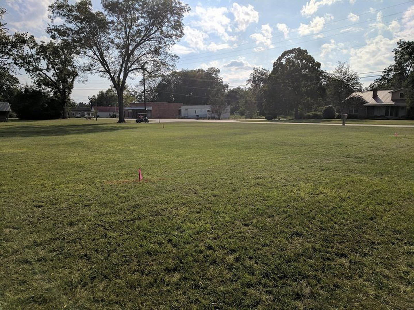 Grassy field bordered by mature trees, single-story house with brick chimney, blue sky with scattered clouds overhead