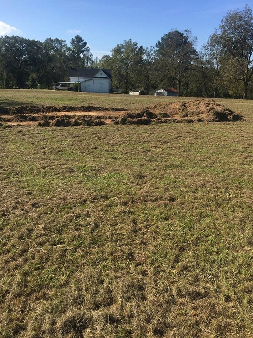 White house with black roof set behind a large grassy field, dirt patches in foreground, trees and blue sky with clouds in background