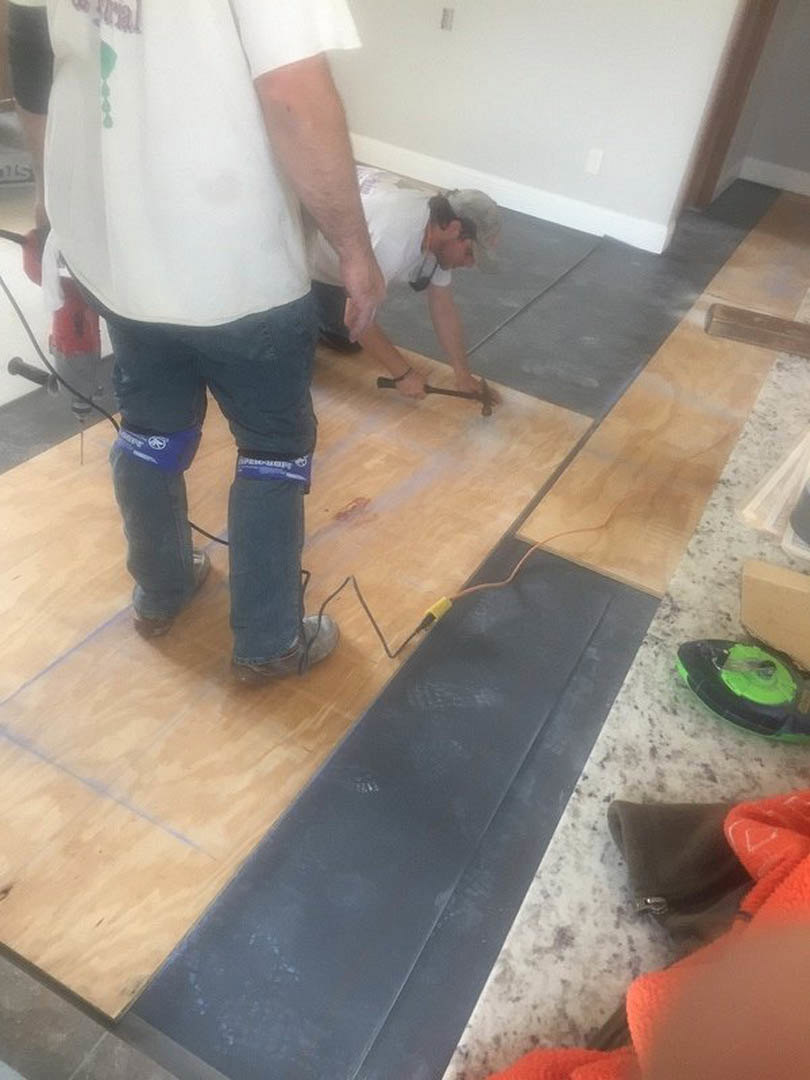 Man in jeans and knee pads bending over unfinished wood floor, using a hammer near a green and black tool, with black mat and shirt visible in indoor residential space.