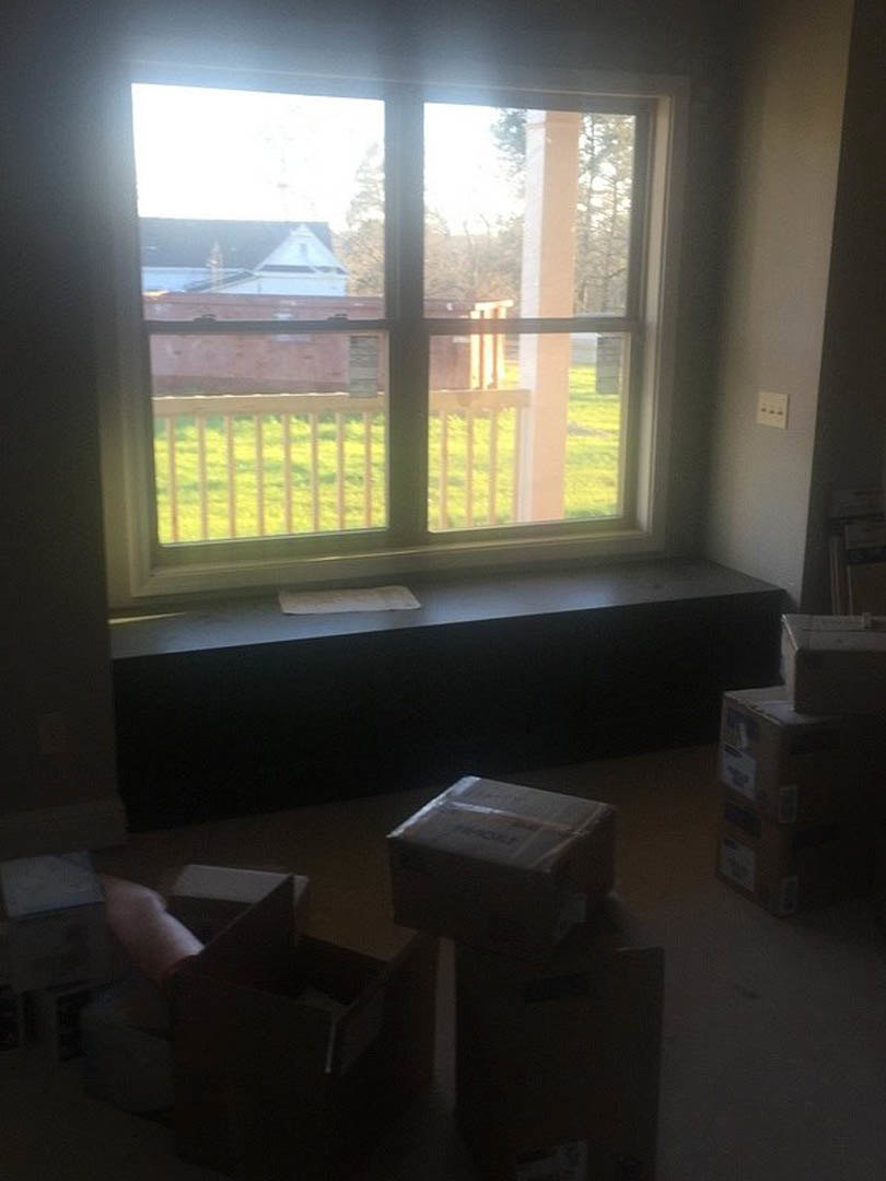 Stacked cardboard boxes on hardwood floor near a large window, white walls, partial view of porch outside, person's arm reaching into a drawer