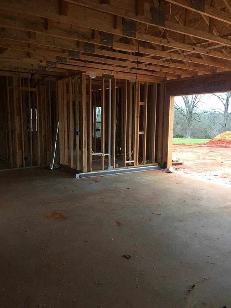 Wood-framed custom home under construction with attached garage, exposed lumber beams, unfinished wooden ceiling, tree visible through window, dirt field and road in foreground