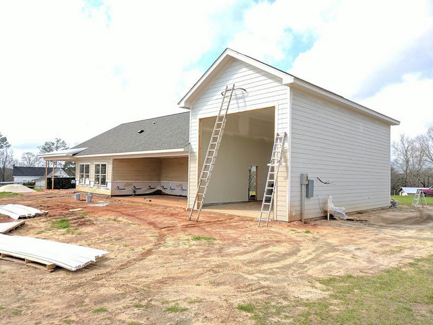 Aluminum ladder leaning against white siding near garage, pallet of construction materials on ground, cloudy sky above, trees in background