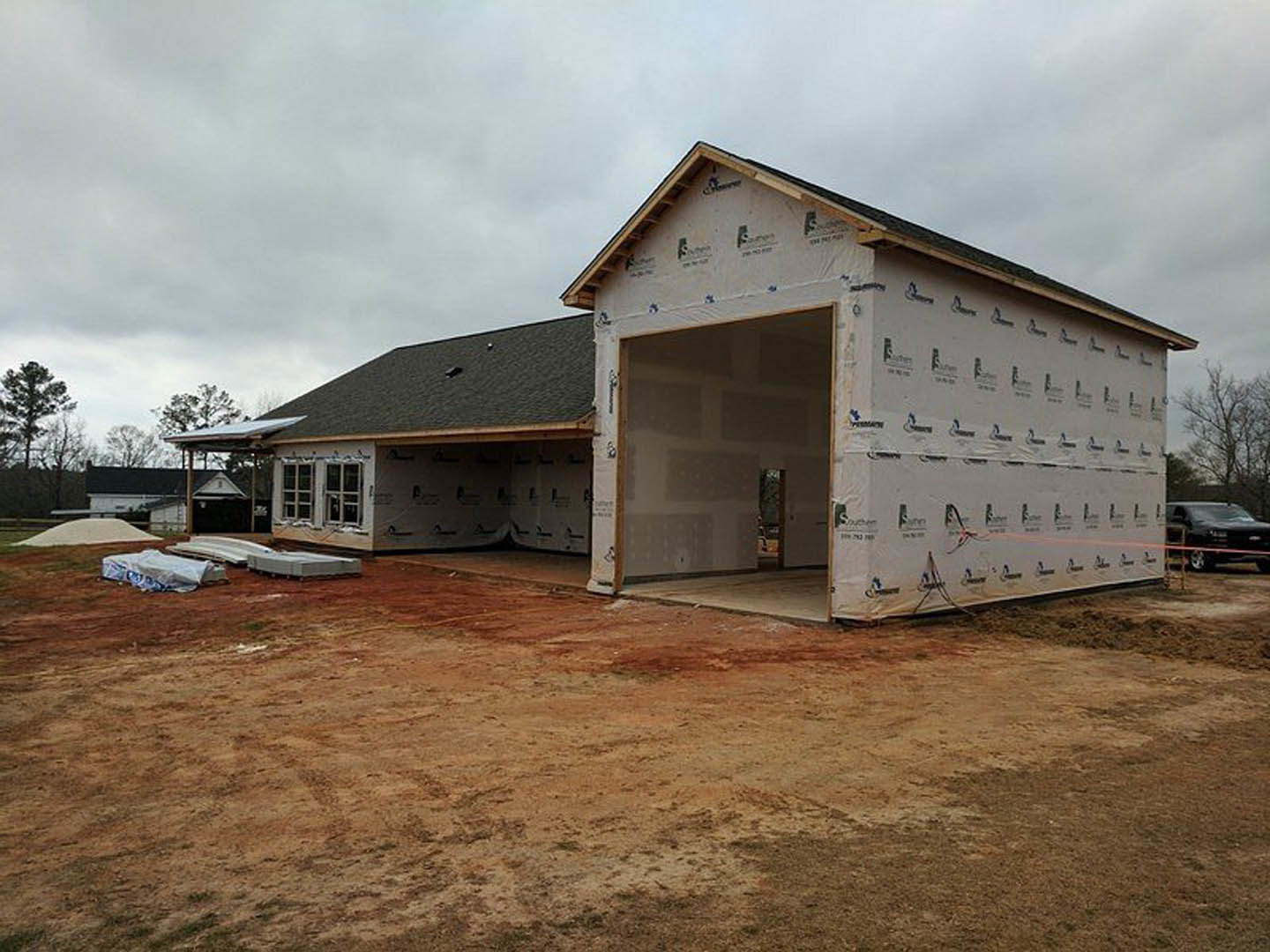 Framed house under construction with exposed plywood, open garage door, dirt lot, scattered white boards, black van parked in driveway, large windows, cloudy sky overhead