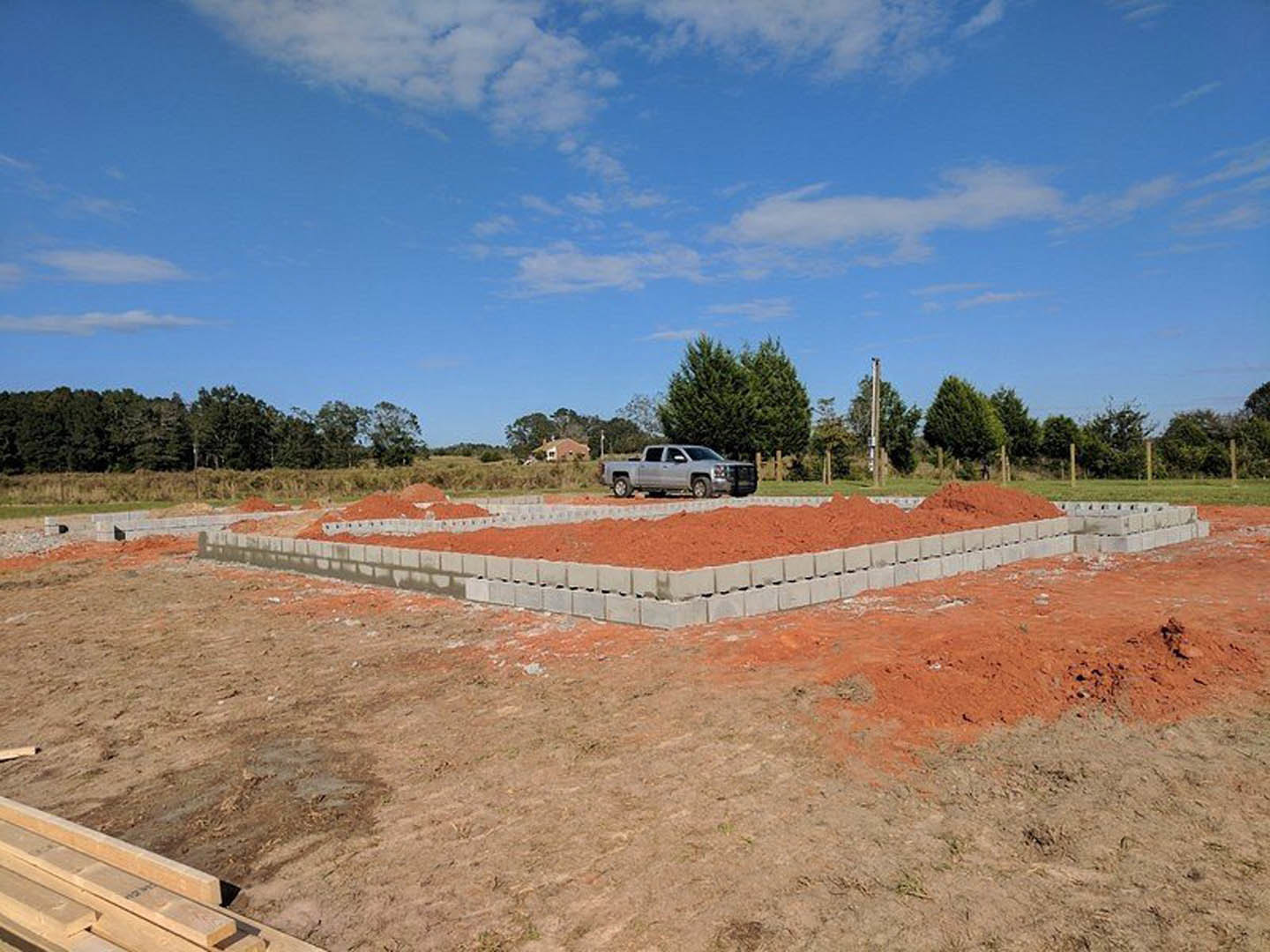 Silver pickup truck parked on red dirt beside concrete block foundation, wood beam in foreground, leafy tree and blue sky with scattered clouds in background