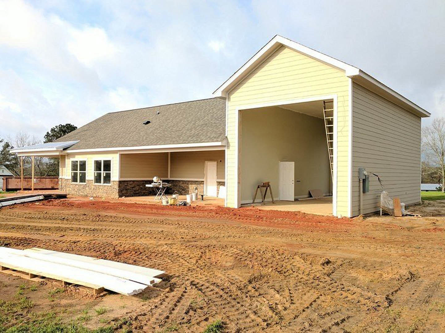 Partially built house with exposed framing, white metal beam resting on wooden pallet, ladder leaning near white door with multiple knobs, dirt road and area marked by tire tracks