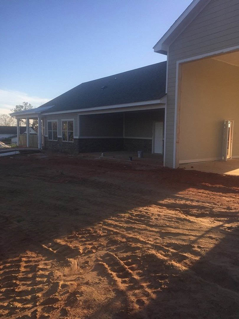 White custom home under construction with dirt yard, visible tire tracks, multi-pane window, exterior door, white wall with side light, and partially finished roof.