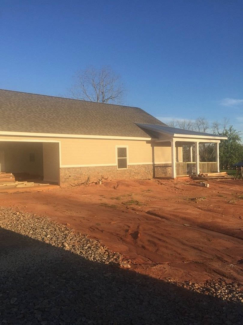 Partially built house with exposed brick walls, square window panes, roof overhang, dirt road leading to gated entrance, tree visible behind roof, construction materials scattered
