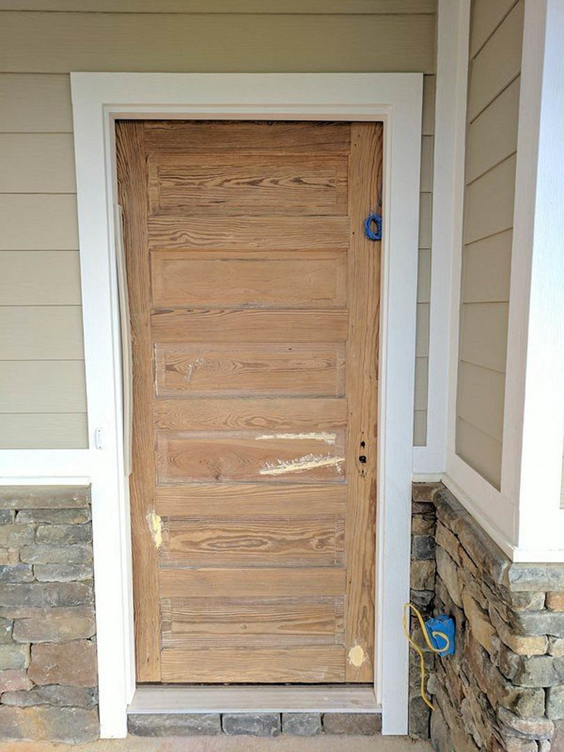 Weathered wooden door with blue handle, surrounded by stone and brick walls, yellow wire resting on wood plank, white wall visible nearby