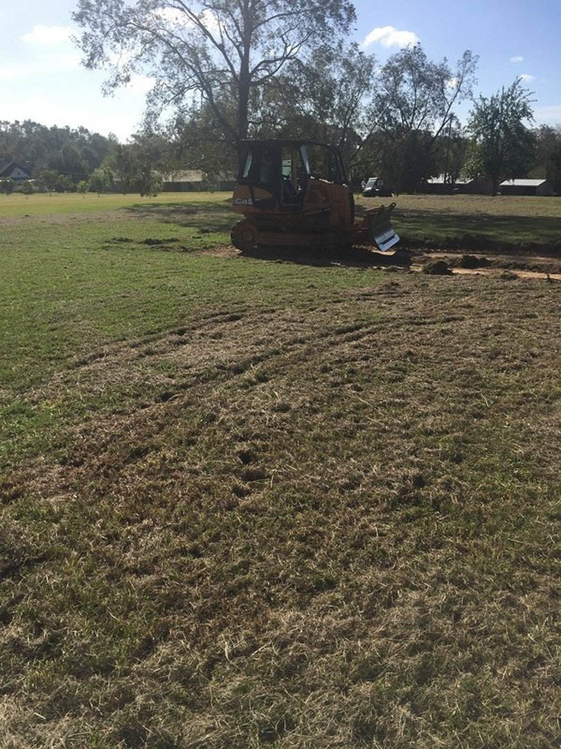 Bulldozer with metal bucket parked on grassy field, Antonine Wall visible in background, blue sky with scattered clouds, leafy tree nearby