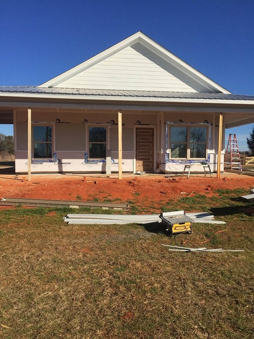 Wood-framed house under construction with exposed beams, ladder leaning against unfinished porch, white-framed window, grassy yard, and clear blue sky