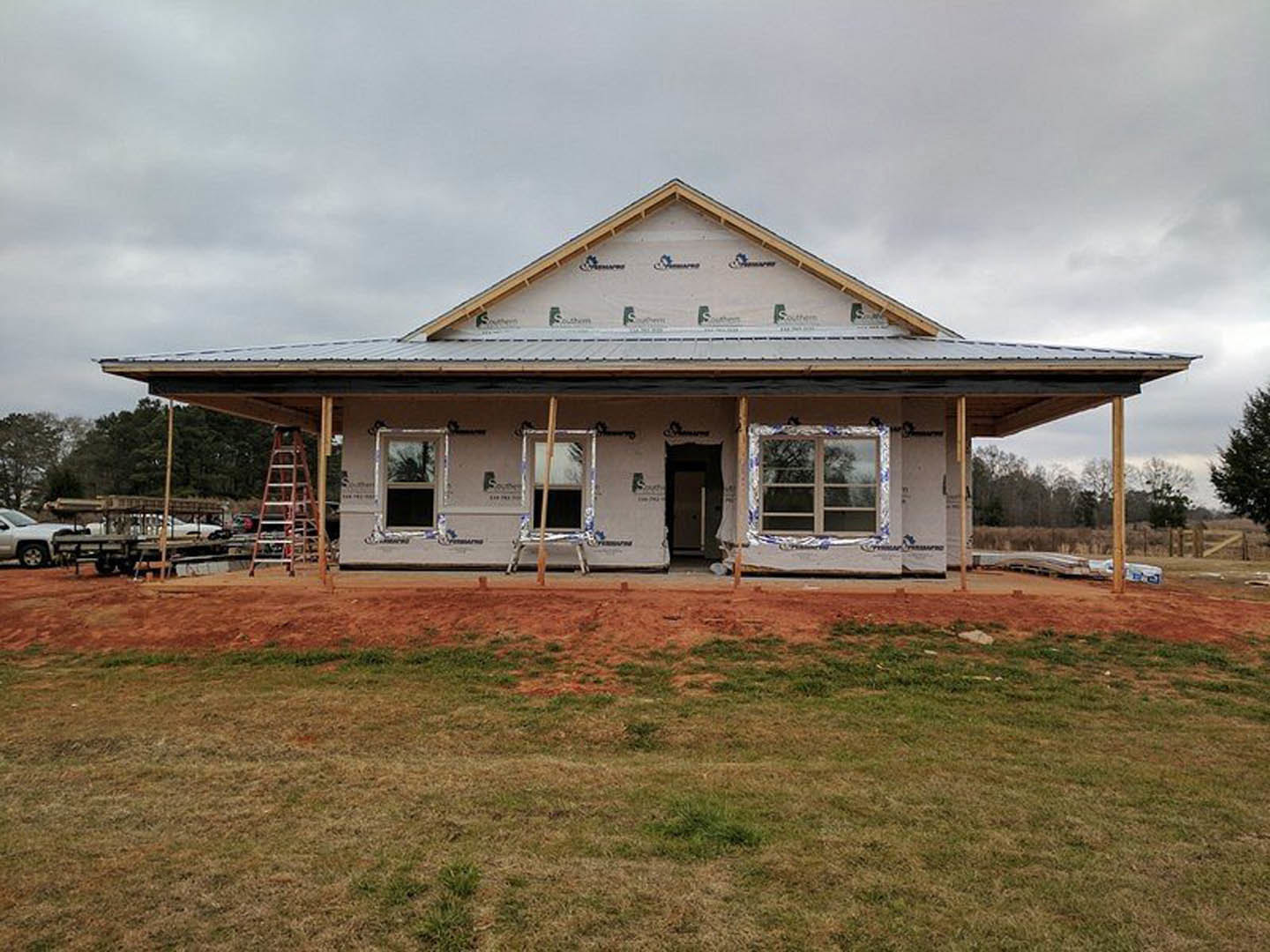 Framed house under construction with exposed wood, ladder leaning against porch, grassy lot, white vehicle parked in background, triangular roof structure, white-framed windows