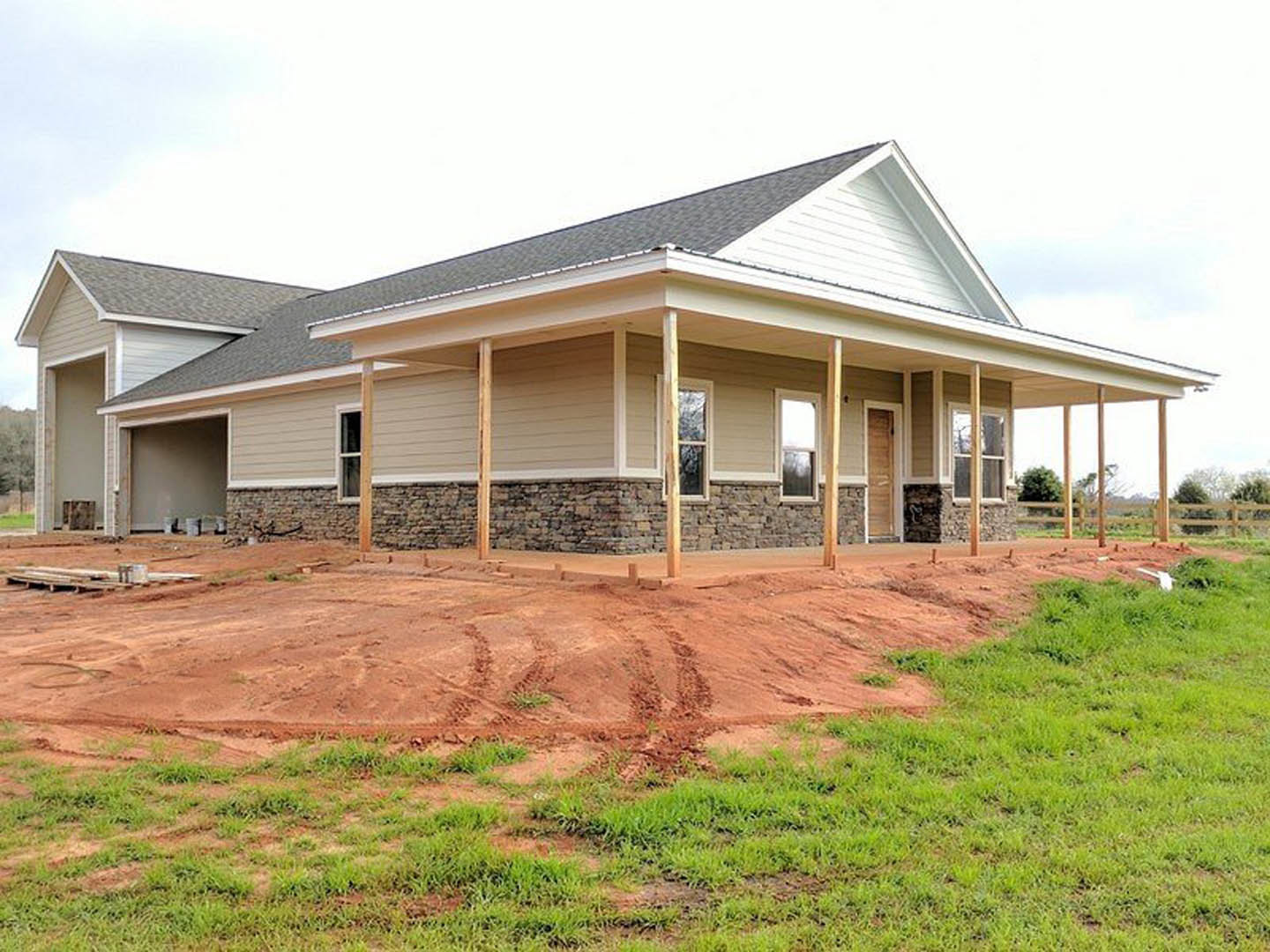 Two-story house under construction with exposed framing, brick walls, and a covered porch, surrounded by a dirt field with tire tracks and patches of grass.
