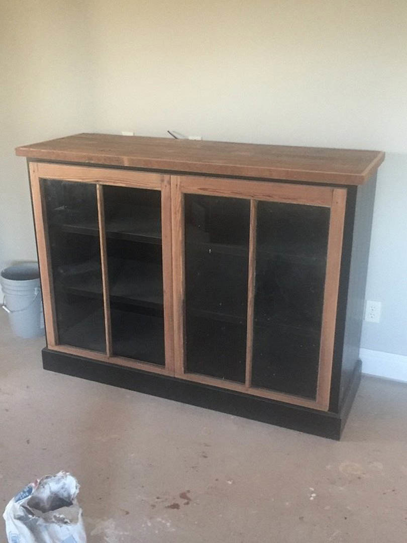 Black and wood cabinet with glass doors, white floor with black trim, torn white object on the floor, wooden surface with black stripe, close-up of a bucket