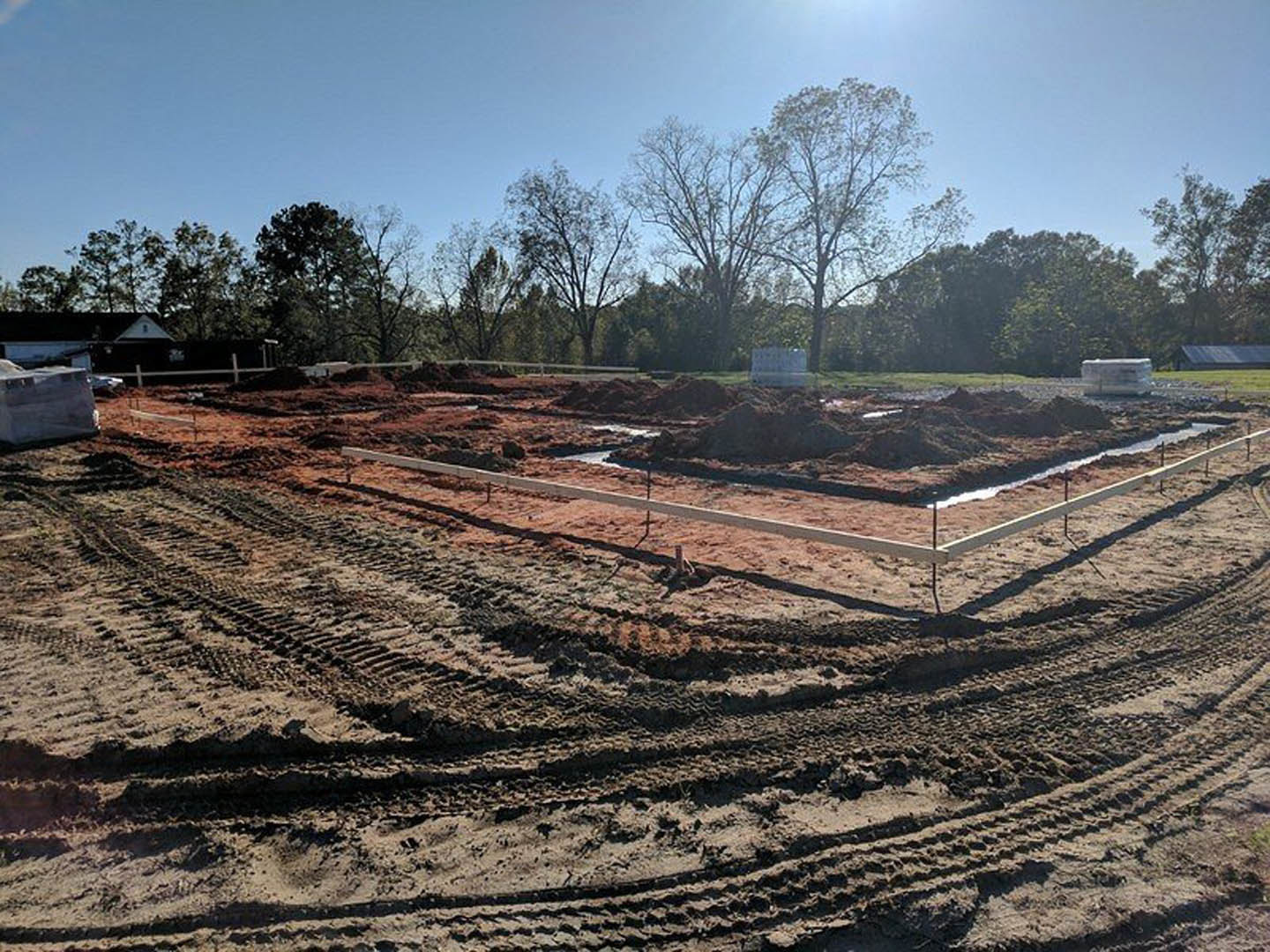 Dirt construction site with tire tracks, scattered beams, trees in the background, blue sky, and a large white box on the ground