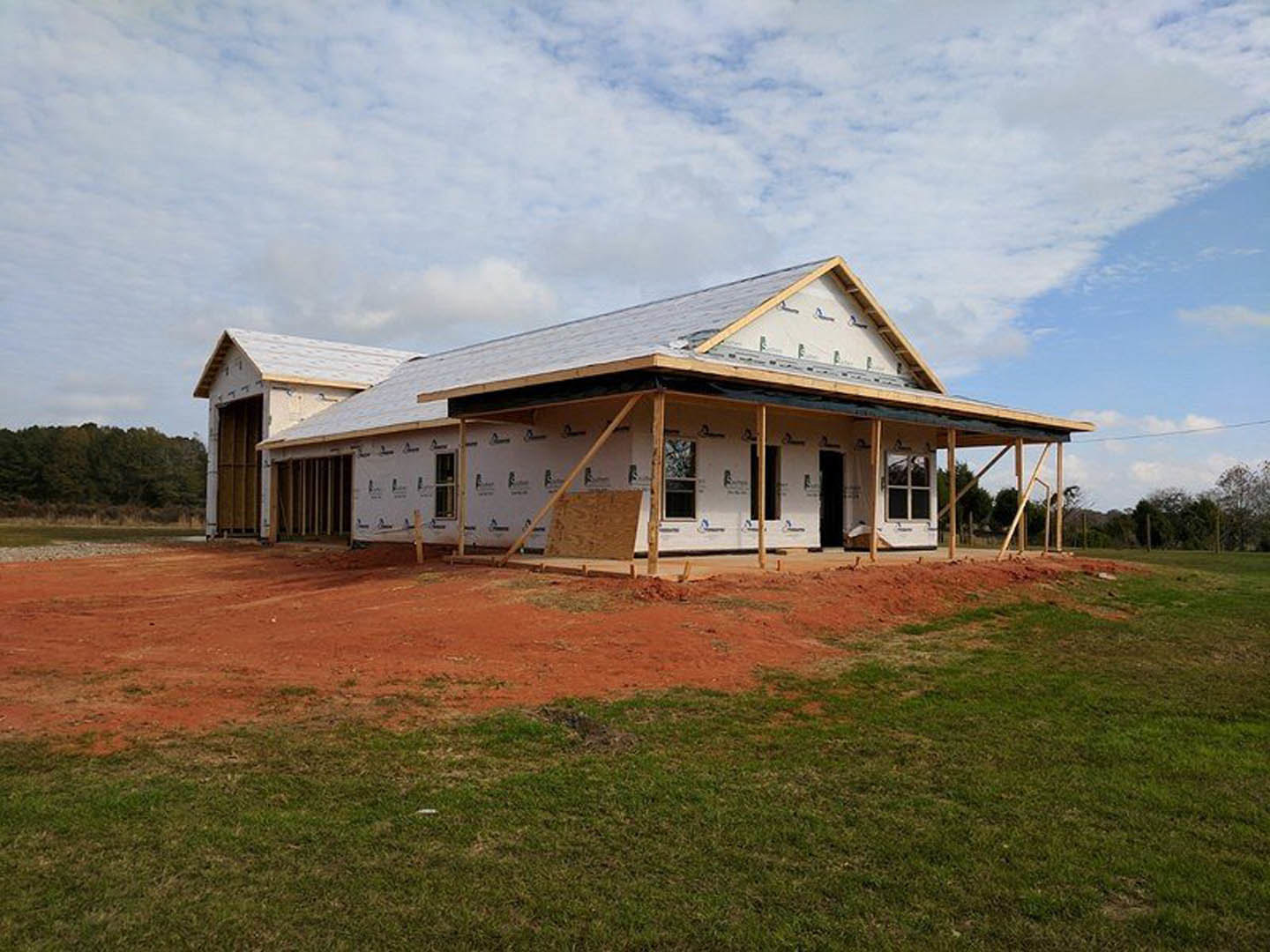 Two-story farmhouse under construction featuring exposed wood beams, expansive covered porch, grassy yard, and surrounding trees under a cloudy sky