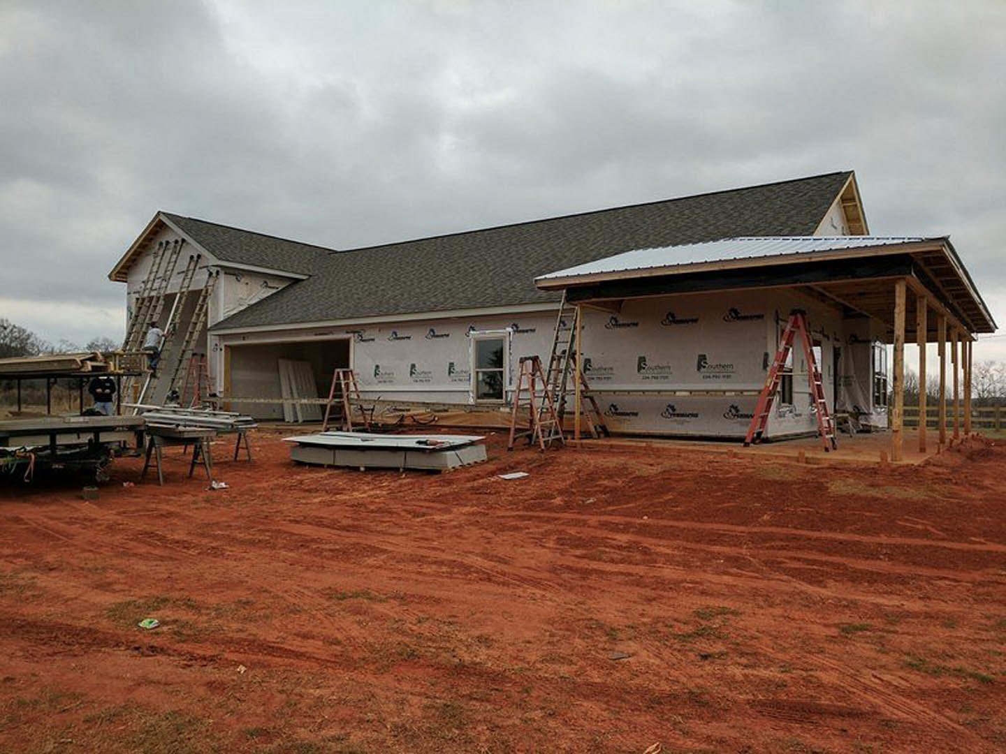 Wood-framed house under construction with exposed sheathing, ladder leaning against exterior wall, dirt yard, and cloudy sky overhead