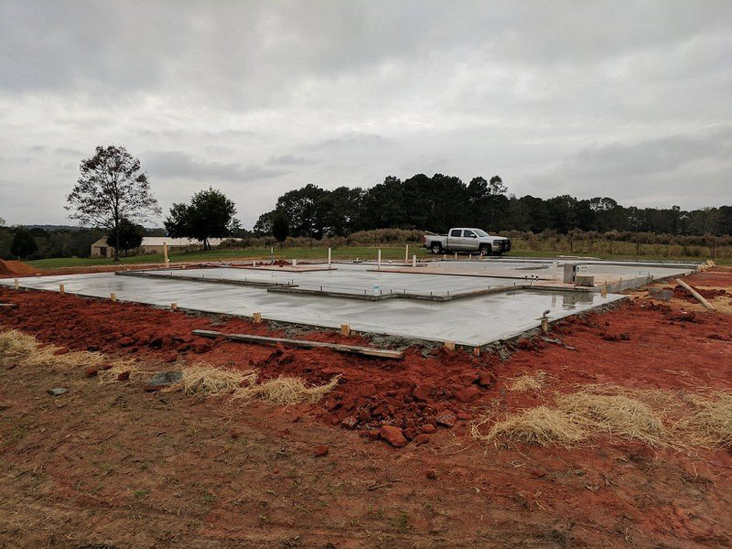 White pickup truck parked on red dirt beside concrete foundation, leafless tree and cluster of trees under cloudy sky, metal rod visible in foreground.