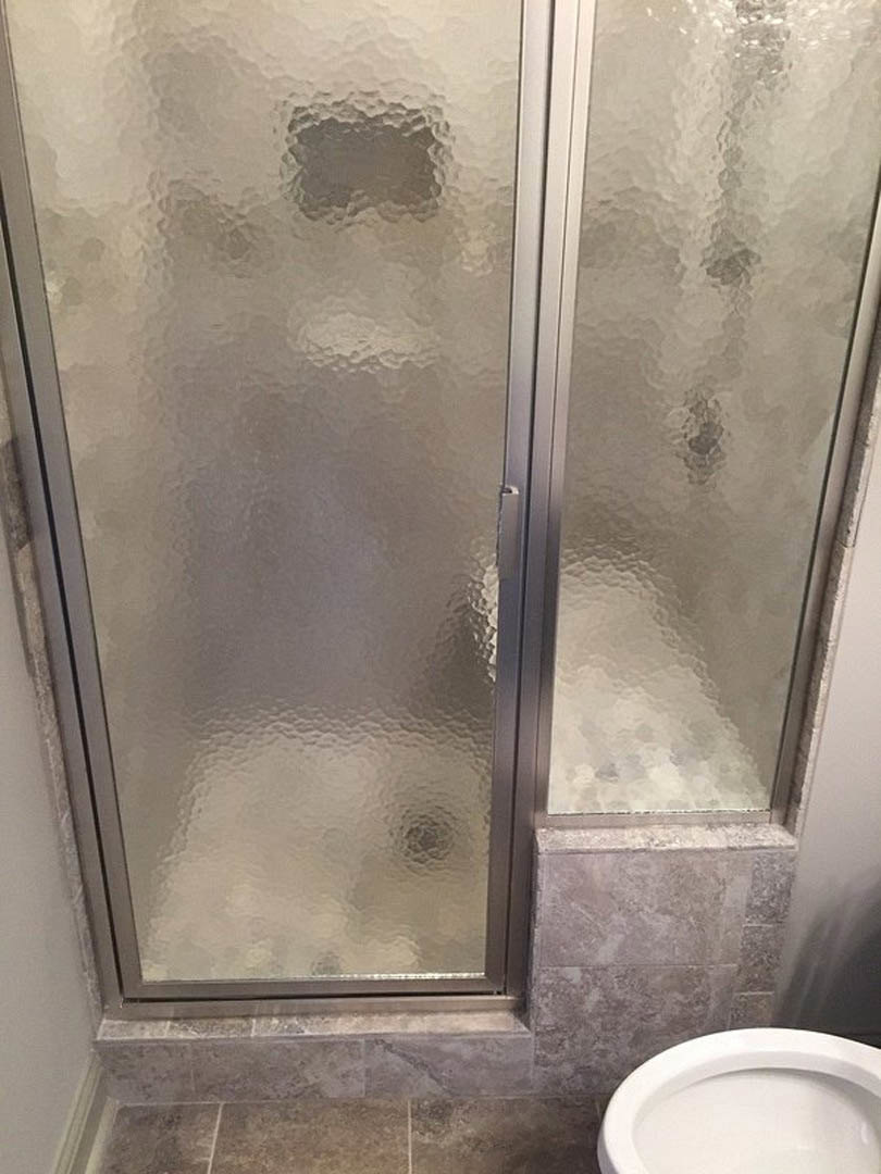 Glass shower door with chrome hardware beside white toilet, gray tile walls, and frosted window in modern bathroom.