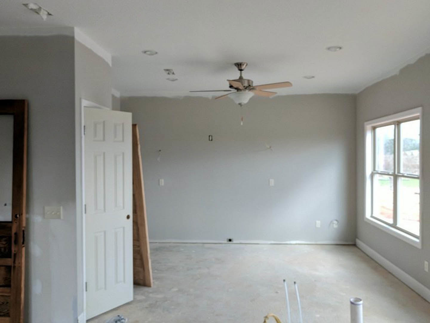 Neutral-toned room with plaster walls, wood flooring, ceiling fan with light fixture, white door with metal doorknob, and window letting in natural light